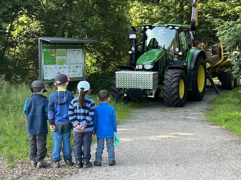 Vier Kinder stehen von hinten am Waldweg und schauen einem Traktor mit Rückewagen zu