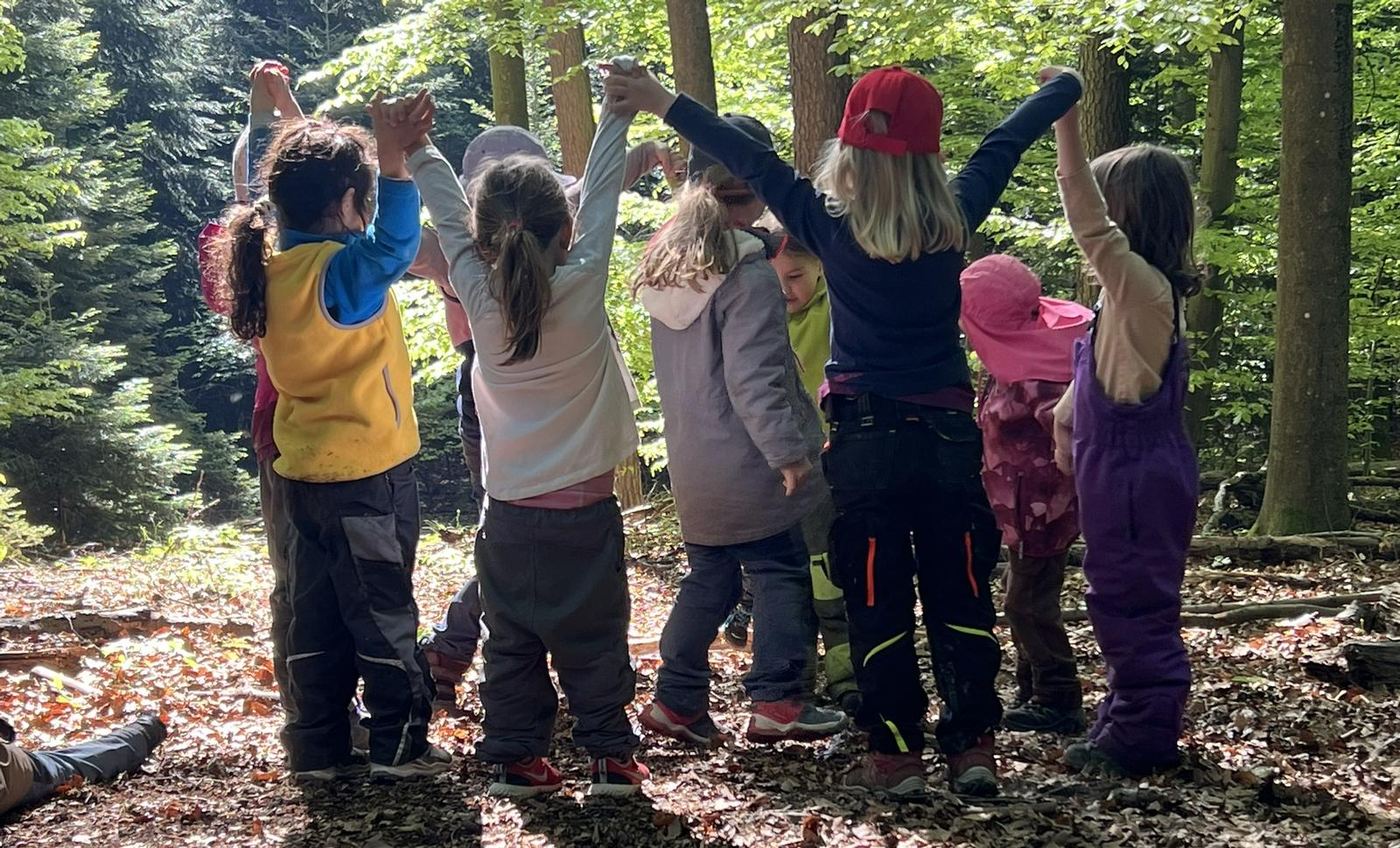 Sommer bei den Waldstrolchen: Kinder stehen im Kreis unter dem Blätterdach und strecken gemeinsam die Arme in die Höhe