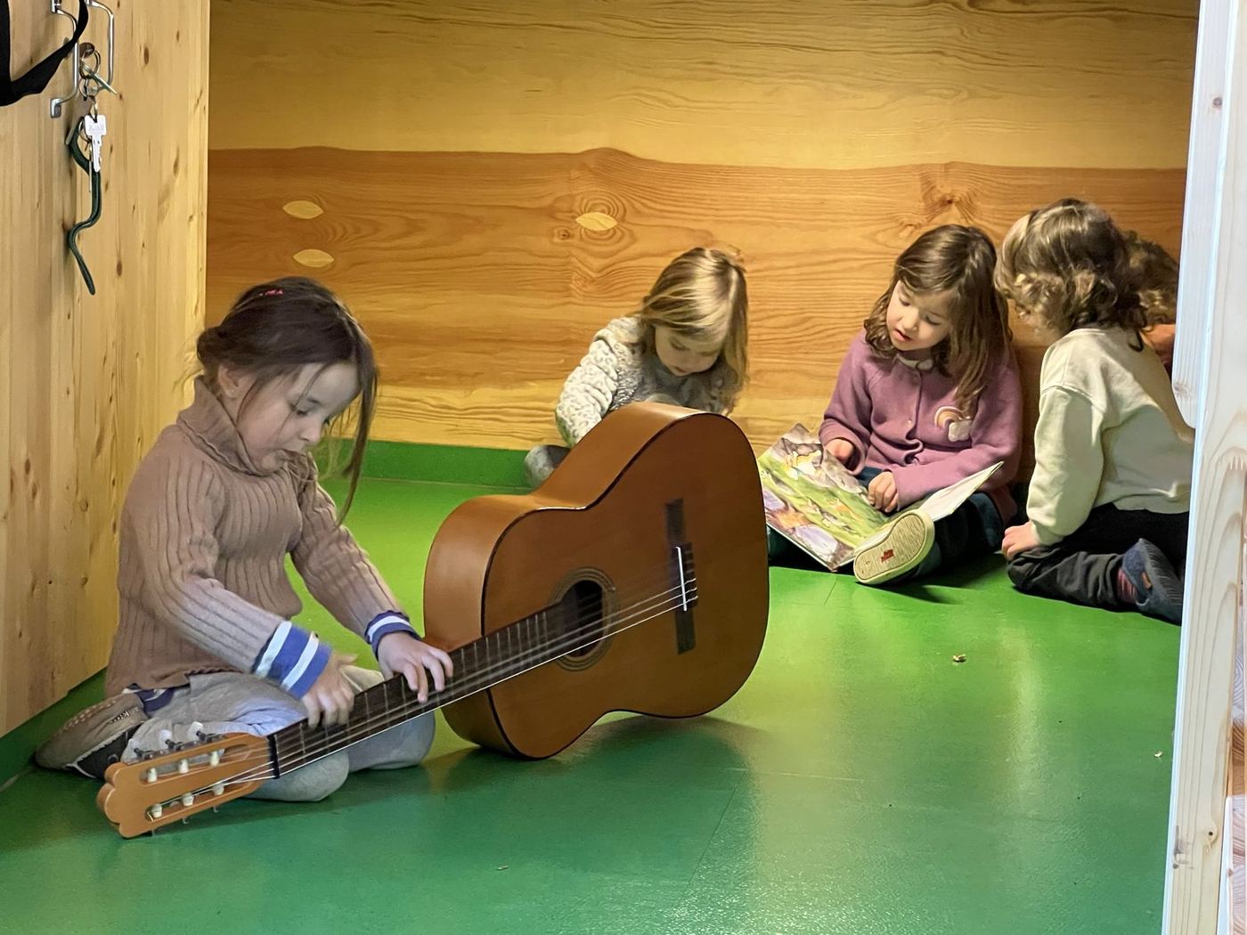 Vier Kinder sitzen im Bauwagen auf dem Boden: eines spielt Gitarre, zwei schauen zusammen ein Bilderbuch an